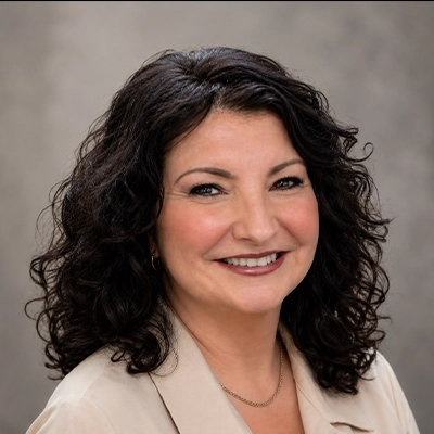Headshot of Alicia with dark curly hair wearing a light-colored blouse against a neutral gray background.
